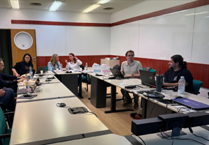 Group of people in a meeting room seated around tables with laptops, discussing and working together during a workshop or project session.